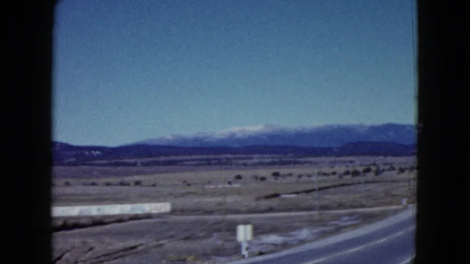 1958 Las Vegas-area road view with a paved shoulder, small roadside signs, open desert, and snow-capped mountains in the distance.