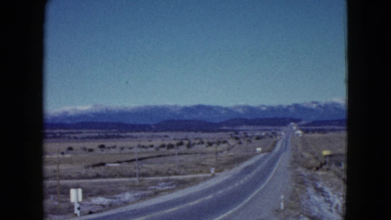 1958 8mm Las Vegas-area road view with a paved highway, desert plain, sparse poles, and mountains on the horizon.