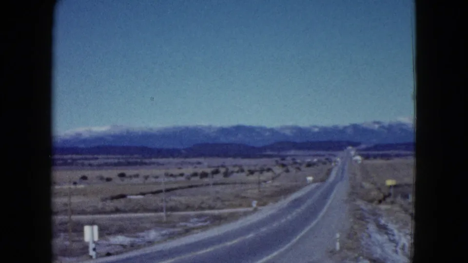 1958 8mm Las Vegas-area road view with a paved highway, desert plain, sparse poles, and mountains on the horizon.