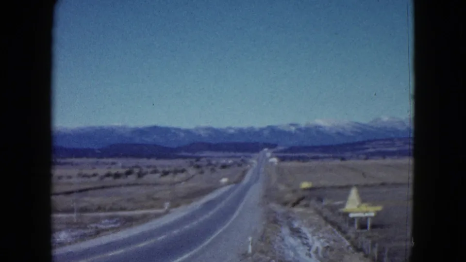 Vehicle-shot 1958 Las Vegas-area highway footage showing open desert, a roadside attraction sign, and snow-capped mountains.