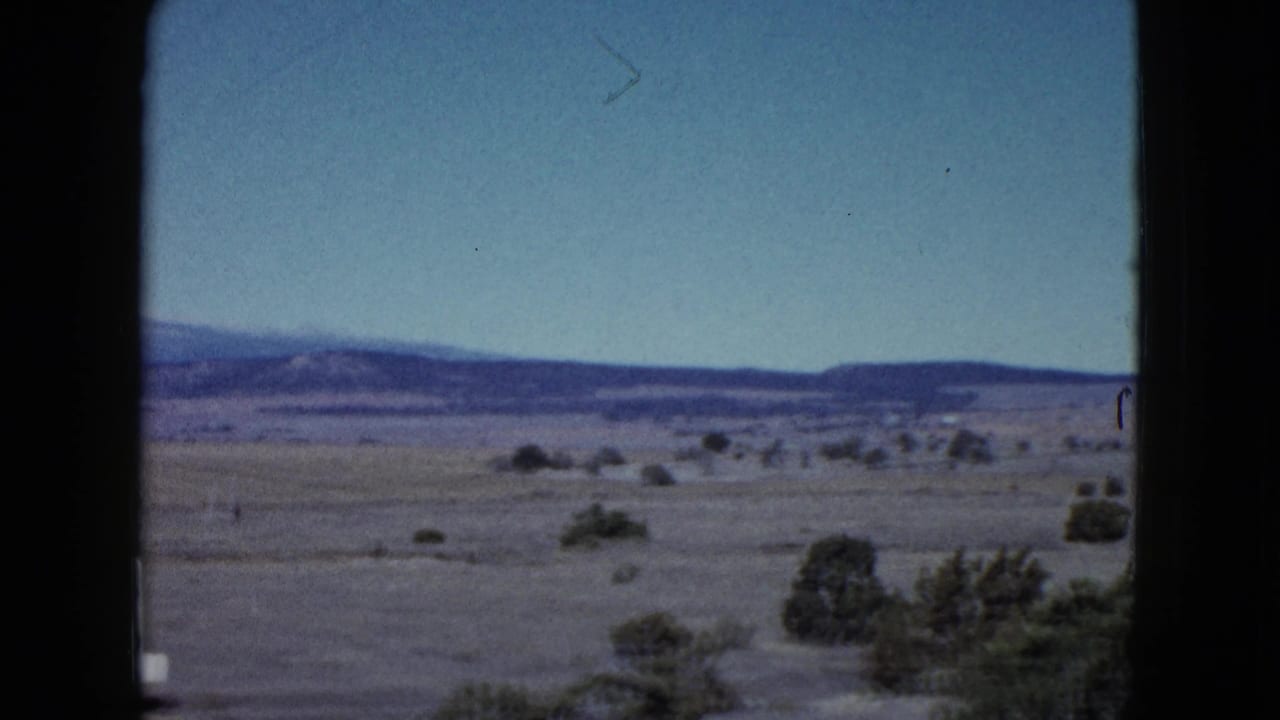 Desert plain with scattered shrubs, distant hills, blue sky, and dark window edges.