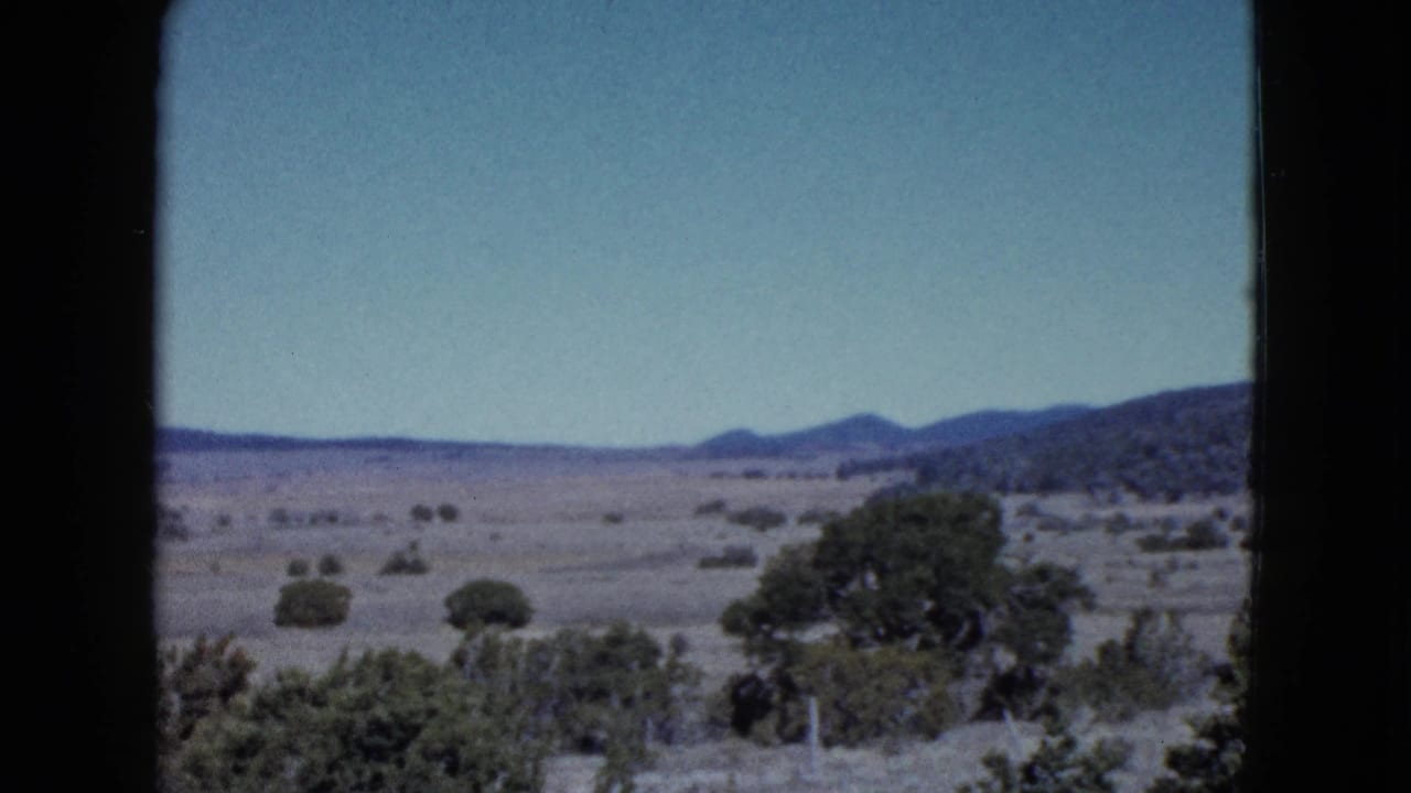 Wide desert landscape with shrubs, distant hills, blue sky, and dark film borders.