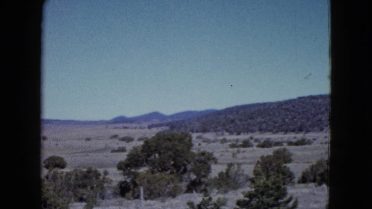 Archival 8mm frame showing a desert vista with scrub vegetation, low hills, pale blue sky, and dark gate edges.