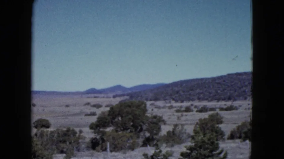 Archival 8mm frame showing a desert vista with scrub vegetation, low hills, pale blue sky, and dark gate edges.