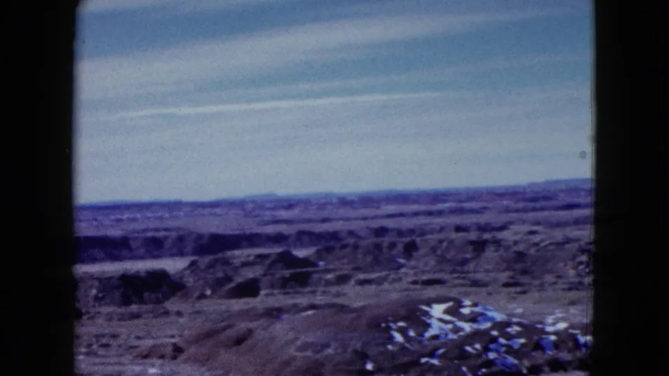 A later frame in the Painted Desert travel shot shows eroded hills, distant plateau lines, and small light patches on the arid ground.