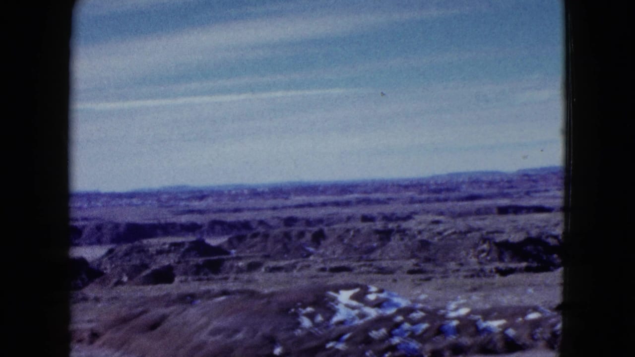 1958 Las Vegas-area home-movie frame showing a barren desert view with eroded ground, distant hills, and a pale blue sky.