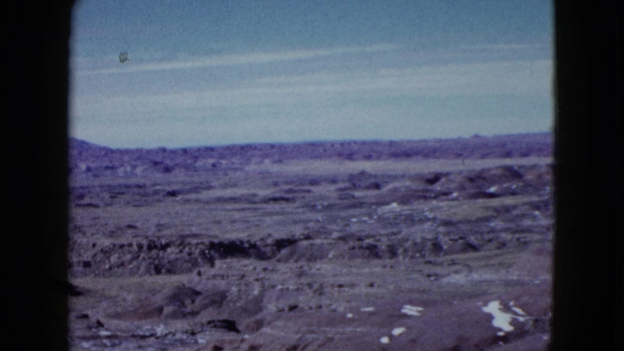 Wide desert basin with eroded ridges, distant hills, and scattered bright ground patches.