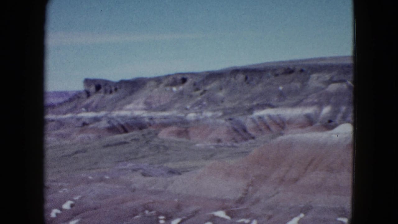 1958 8mm frame from a slow pan across desert badlands near Las Vegas, showing gray and reddish rock layers beneath open sky.