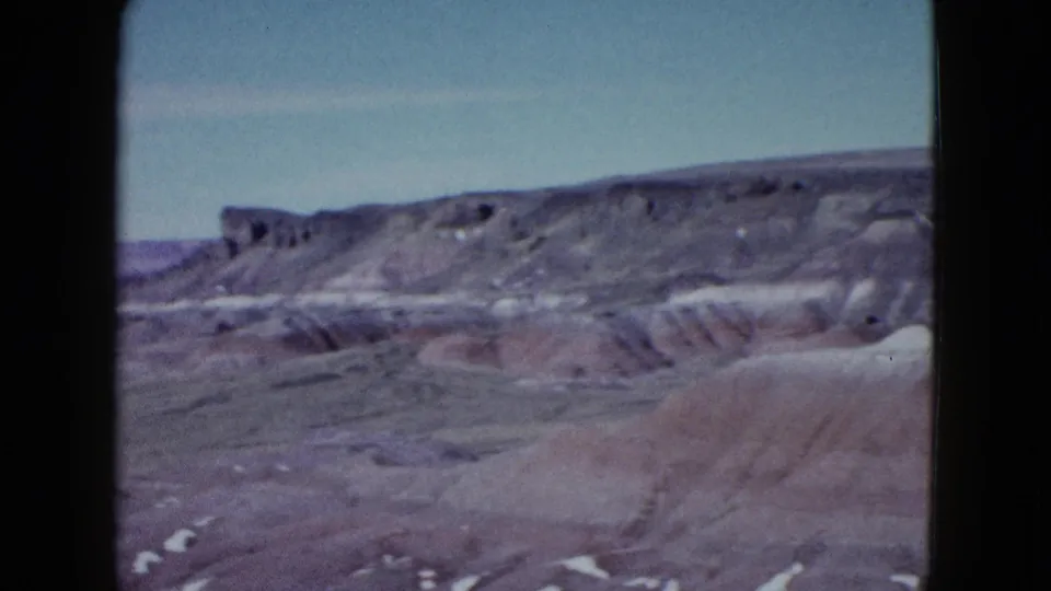 1958 8mm frame from a slow pan across desert badlands near Las Vegas, showing gray and reddish rock layers beneath open sky.