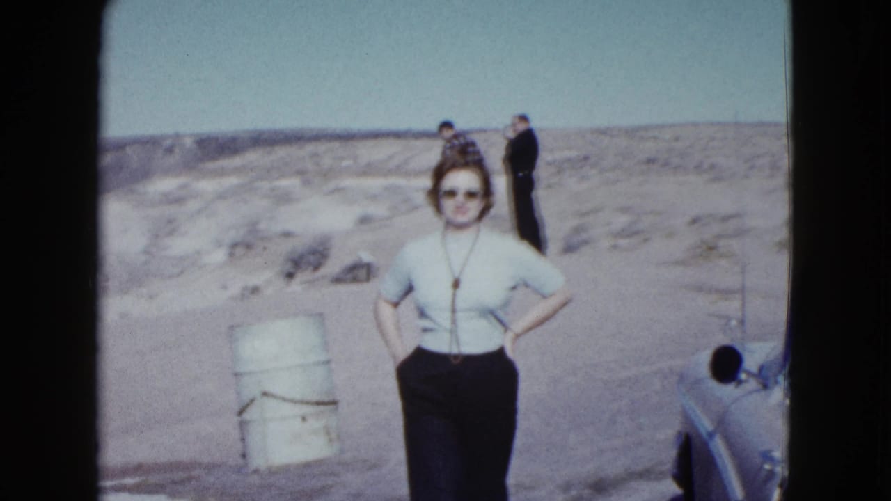 Woman in sunglasses poses in a desert pullout with two men behind her and a partial car at right.
