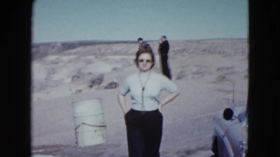 In 1958 Las Vegas-area desert footage, a woman poses near a roadside barrel and partial classic car while two men stand on the ridge behind her.