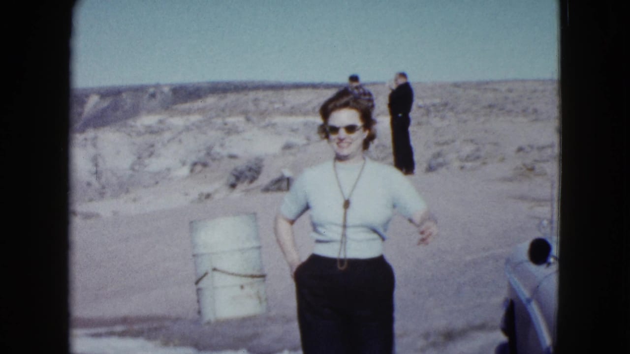 Woman smiles and gestures toward the camera near a barrel, car edge, and two men on desert ground.