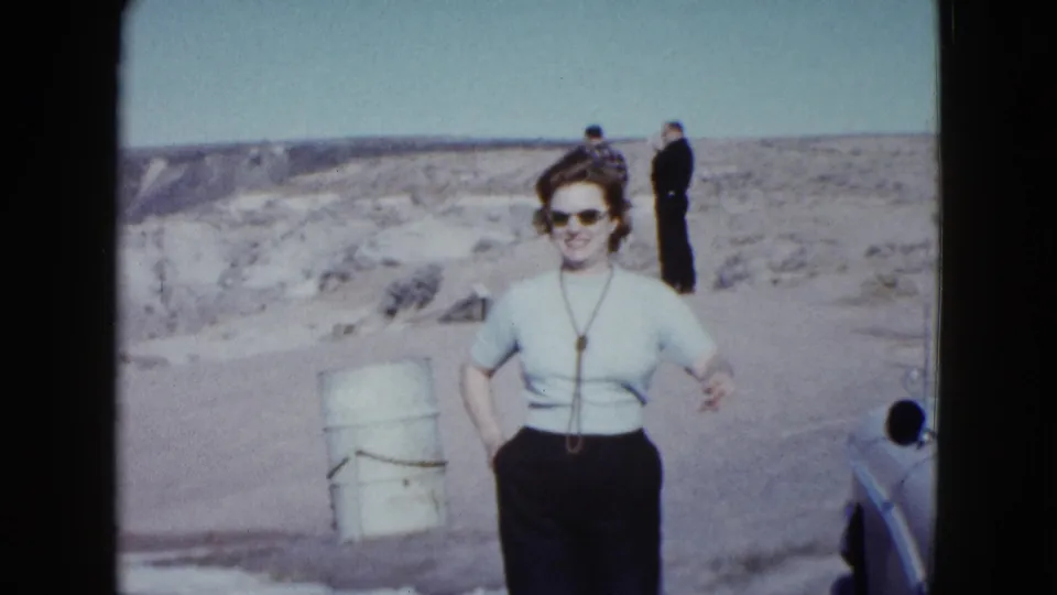 A woman in sunglasses stands in the Las Vegas-area desert, with a white barrel, partial car, and two men visible behind her.