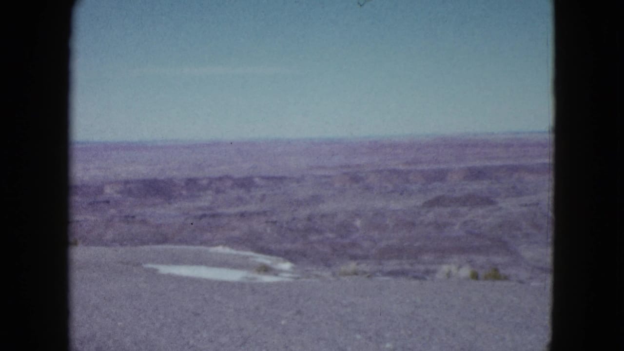 1958 Las Vegas-area home-movie frame showing distant desert ridges, open sky, gravel foreground, and a small pale patch near the ground.