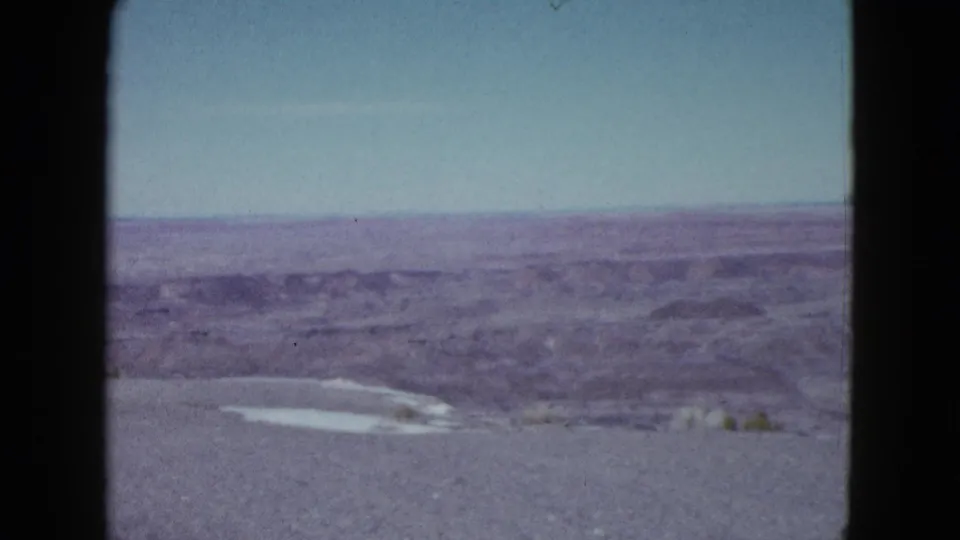 1958 Las Vegas-area home-movie frame showing distant desert ridges, open sky, gravel foreground, and a small pale patch near the ground.