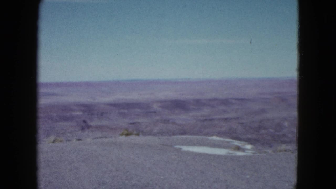 Open desert terrain with distant ridges, pale sky, gravel foreground, and dark frame edge.