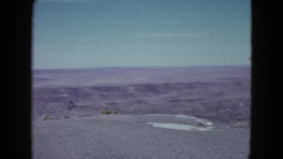1958 Las Vegas-area 8mm frame showing arid terrain, layered ridges, sparse brush, and a pale patch on the foreground ground.