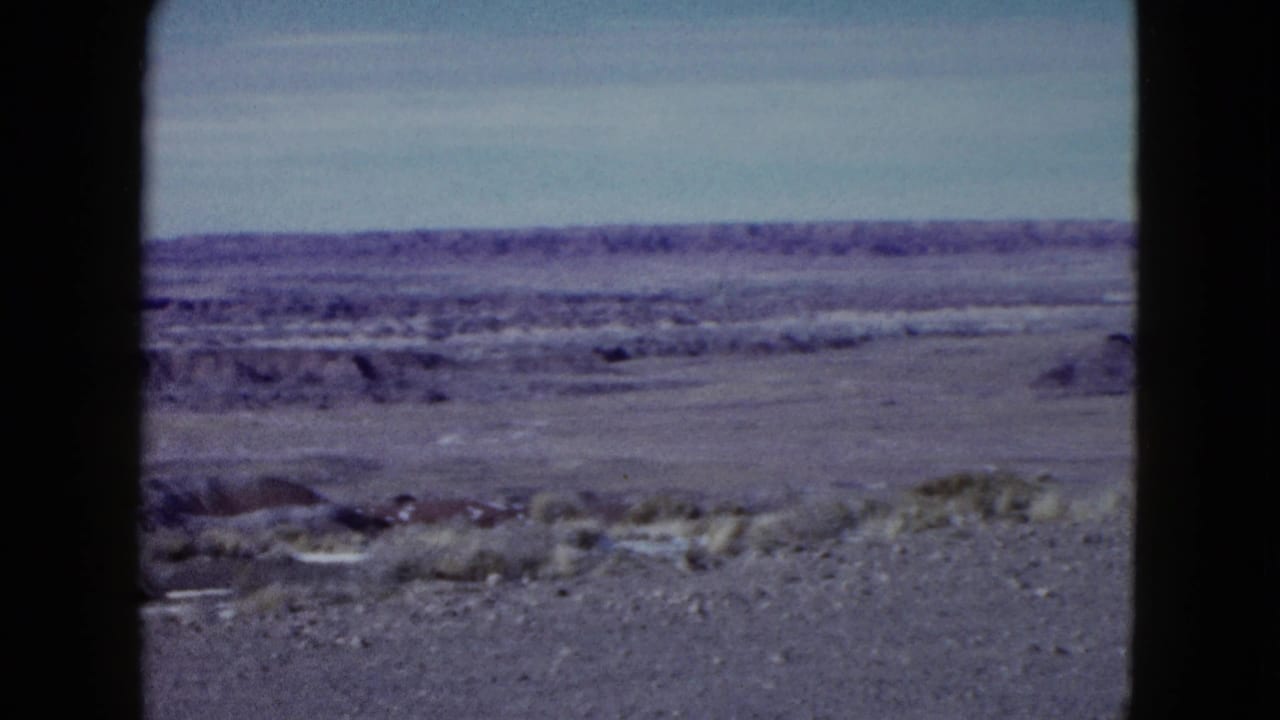 1958 Las Vegas-area home-movie frame showing open desert ground, scrub, pale sky, and distant ridges through dark frame edges.
