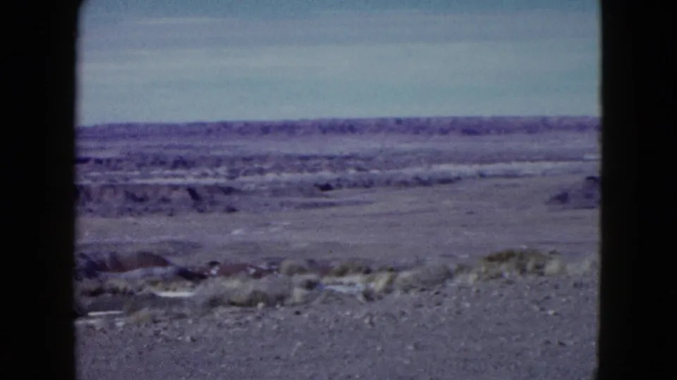 1958 Las Vegas-area home-movie frame showing open desert ground, scrub, pale sky, and distant ridges through dark frame edges.