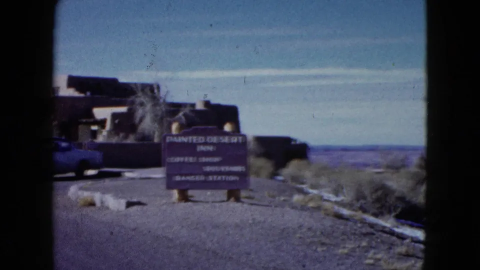 Ground-level 1958 travel view showing a Painted Desert Inn sign, adobe-style buildings, roadside scrub, and blue sky.