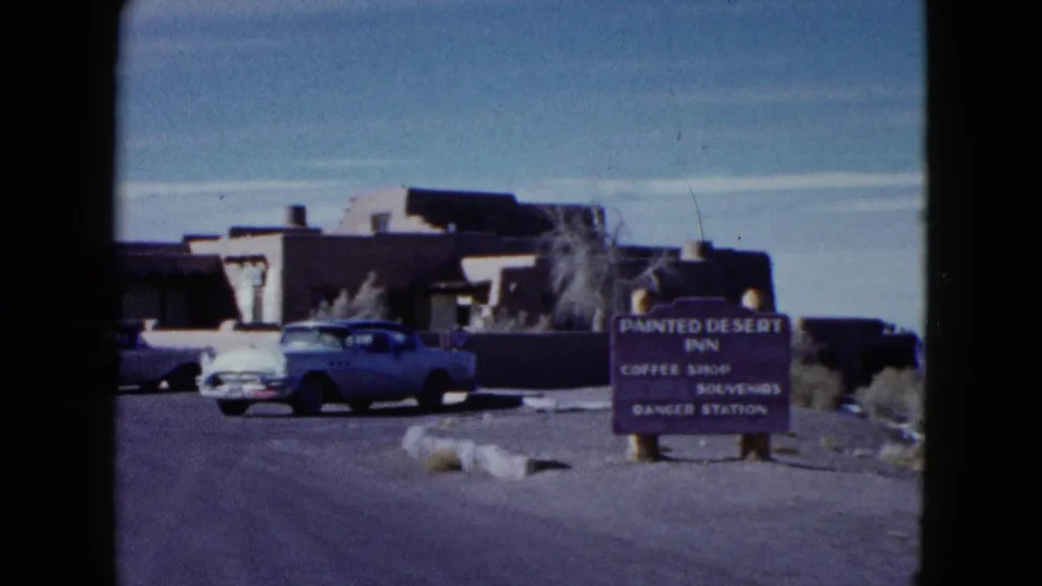 1958 ground-level view of the Painted Desert Inn sign, parked mid-century cars, low adobe-style buildings, and desert roadside vegetation.