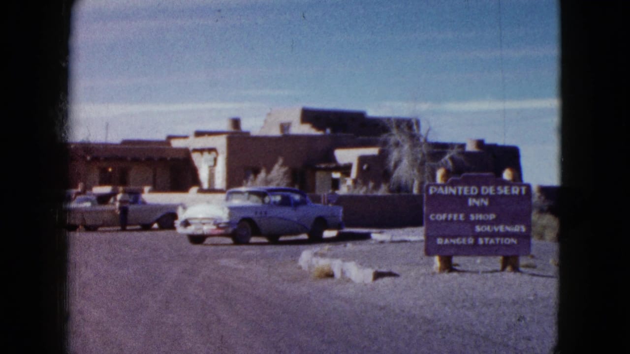 1958 home-movie frame of the Painted Desert Inn exterior with a sign, classic cars, desert landscaping, and adobe-style architecture.