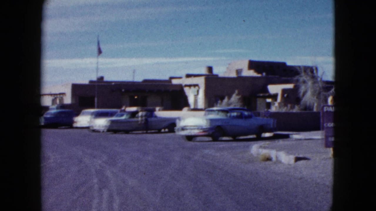 Visitor-shot 1958 frame looking toward the inn approach, with mid-century cars, a flagpole, and the desert building exterior.