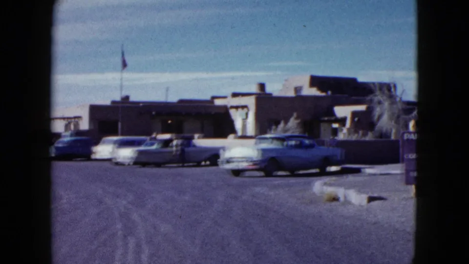 Visitor-shot 1958 frame looking toward the inn approach, with mid-century cars, a flagpole, and the desert building exterior.