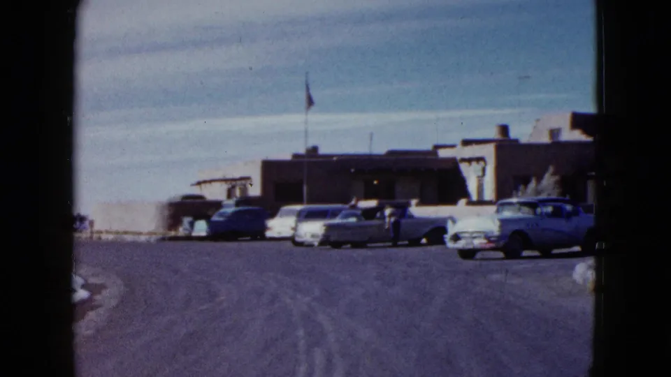 1958 travel footage frame from a moving or handheld viewpoint approaching parked cars near the Painted Desert Inn exterior.