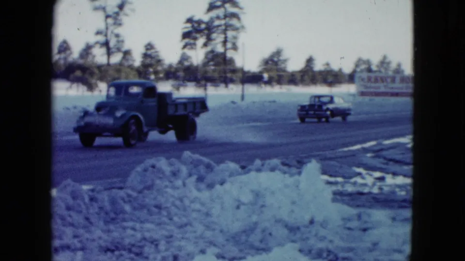 A classic truck drives past snowbanks on a Las Vegas roadside, with another car and the Ranch House sign visible beyond the road.