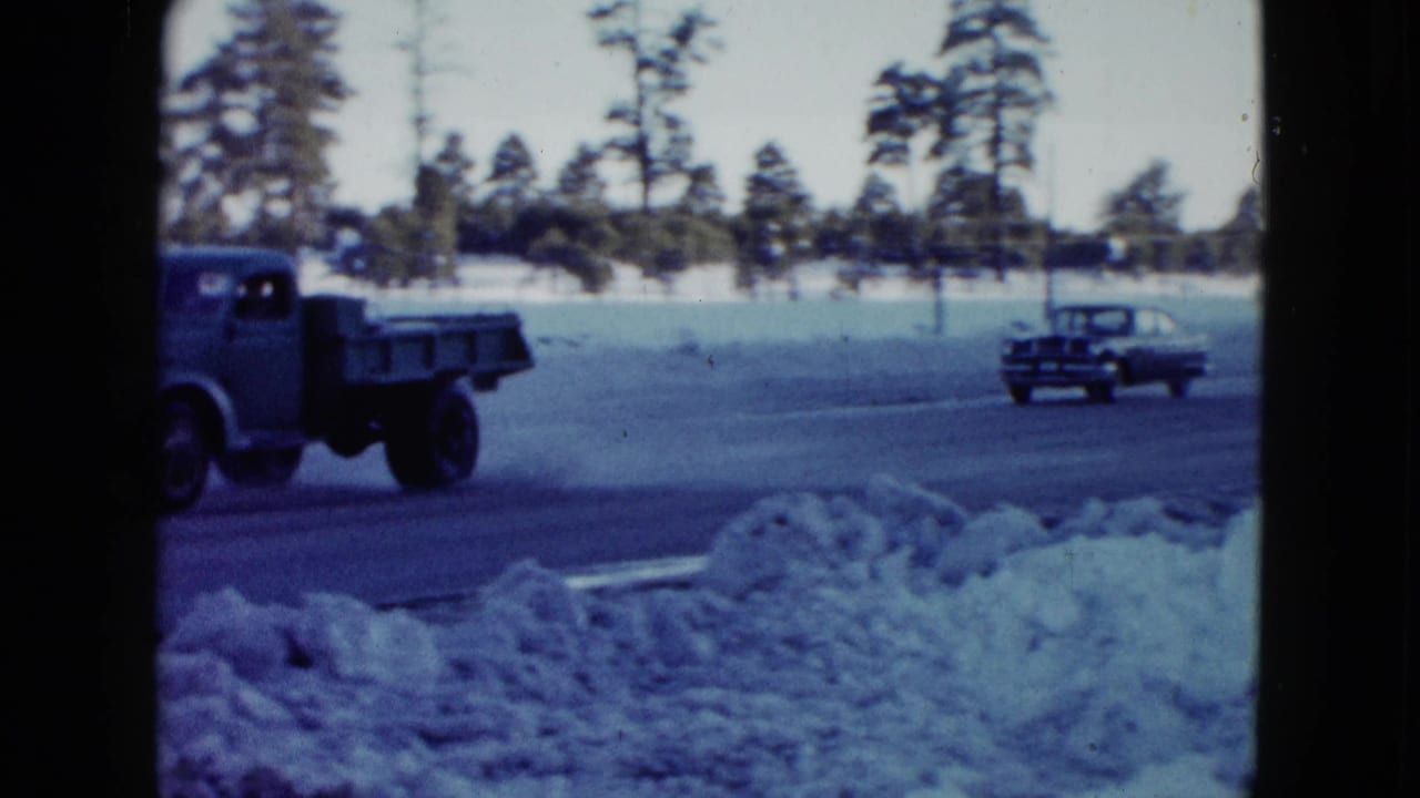 1958 Las Vegas-area 8mm frame showing a vintage truck and car passing snowbanks with tall trees in the background.