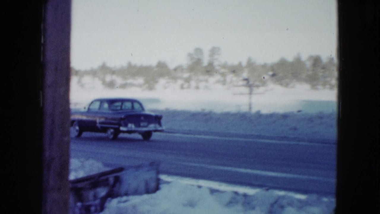 Dark mid-century sedan driving along a wet road bordered by deep snow and distant trees.