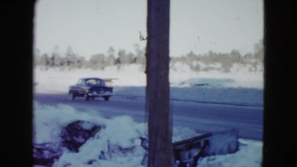 The sedan recedes along a wet snow-lined road, framed by a wooden post in a 1958 Las Vegas-area home movie.