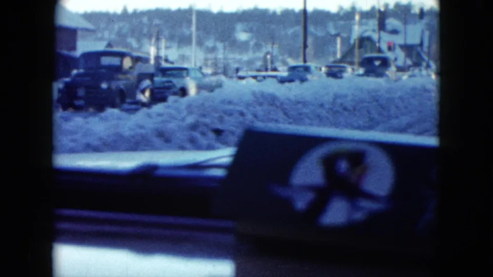 Classic cars line and cross a snowy Las Vegas street in 1958, viewed through the windshield with dashboard and wiper frame visible.