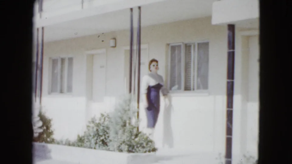 Woman in late-1950s-style clothing standing by a Las Vegas house porch with windows, posts, and low shrubs visible.