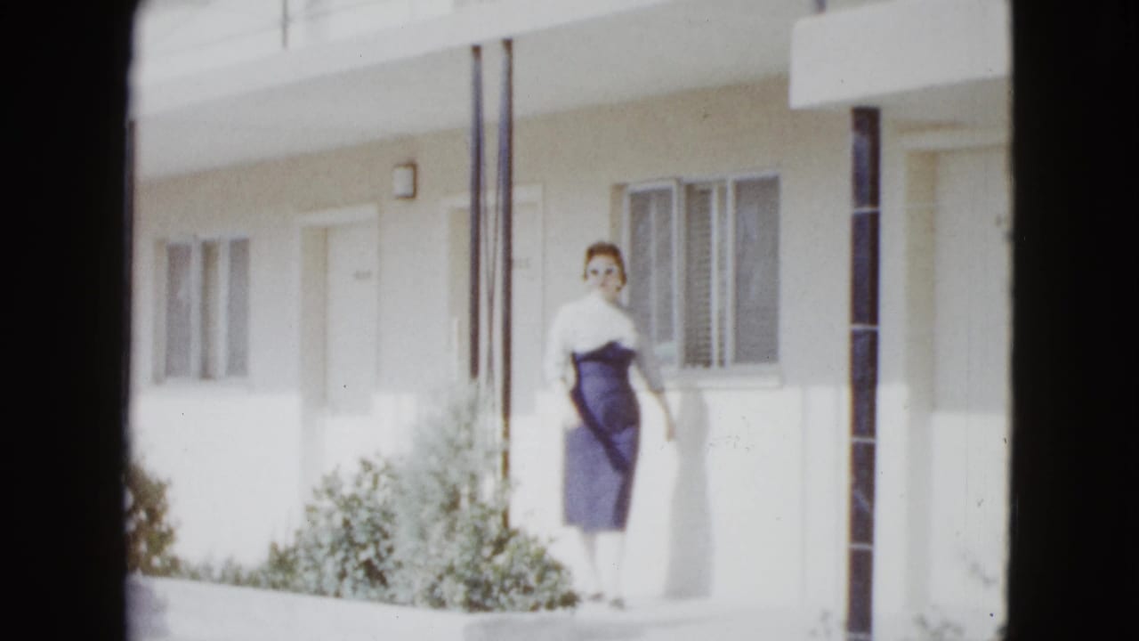 Woman in a fitted dress walks outside a motel room beside windows and shrubs.