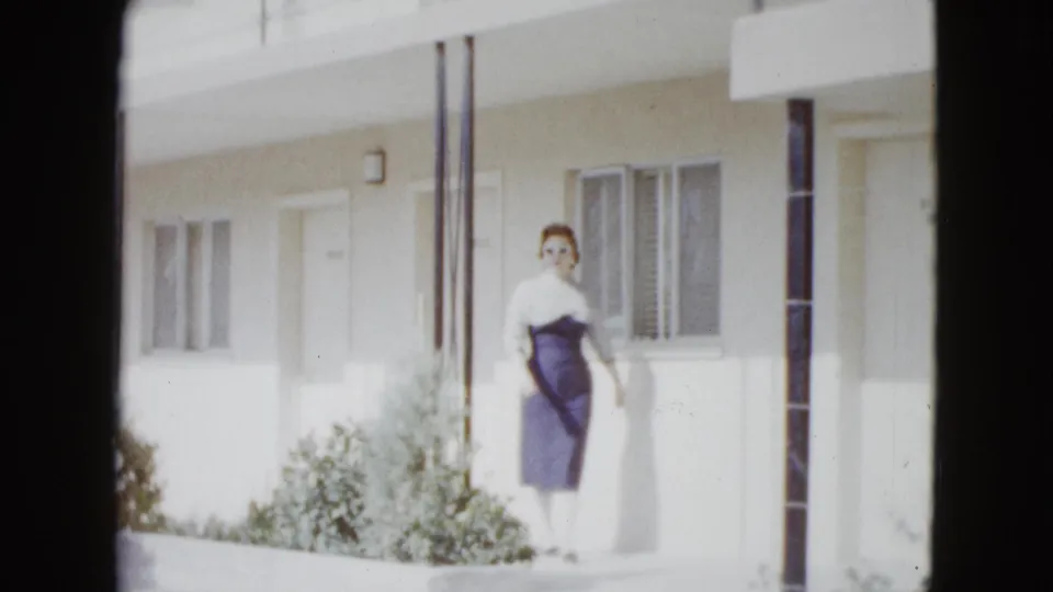 In 1958 Las Vegas footage, a woman in a fitted two-tone dress walks along a bright motel exterior with doors, windows, columns, and shrubs.