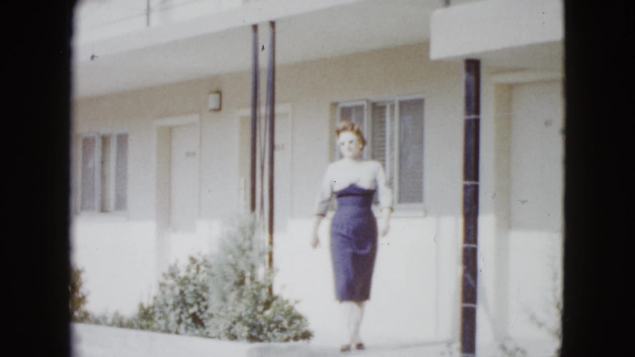 The woman walks toward the camera outside a Las Vegas motel, with white walls, windows, a door, columns, and shrubs behind her.