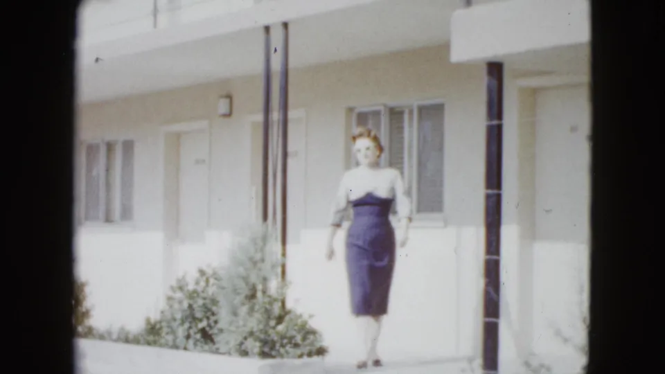 The woman walks toward the camera outside a Las Vegas motel, with white walls, windows, a door, columns, and shrubs behind her.