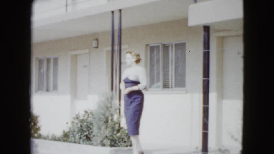 A woman in a dark skirt and light top pauses or turns on a sunlit motel walkway in 1958 Las Vegas home-movie footage.