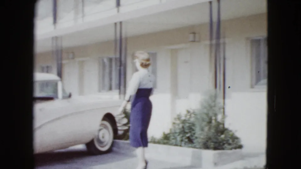 The woman walks away along the motel exterior as a 1950s American car sits parked nearby in the Las Vegas travel scene.