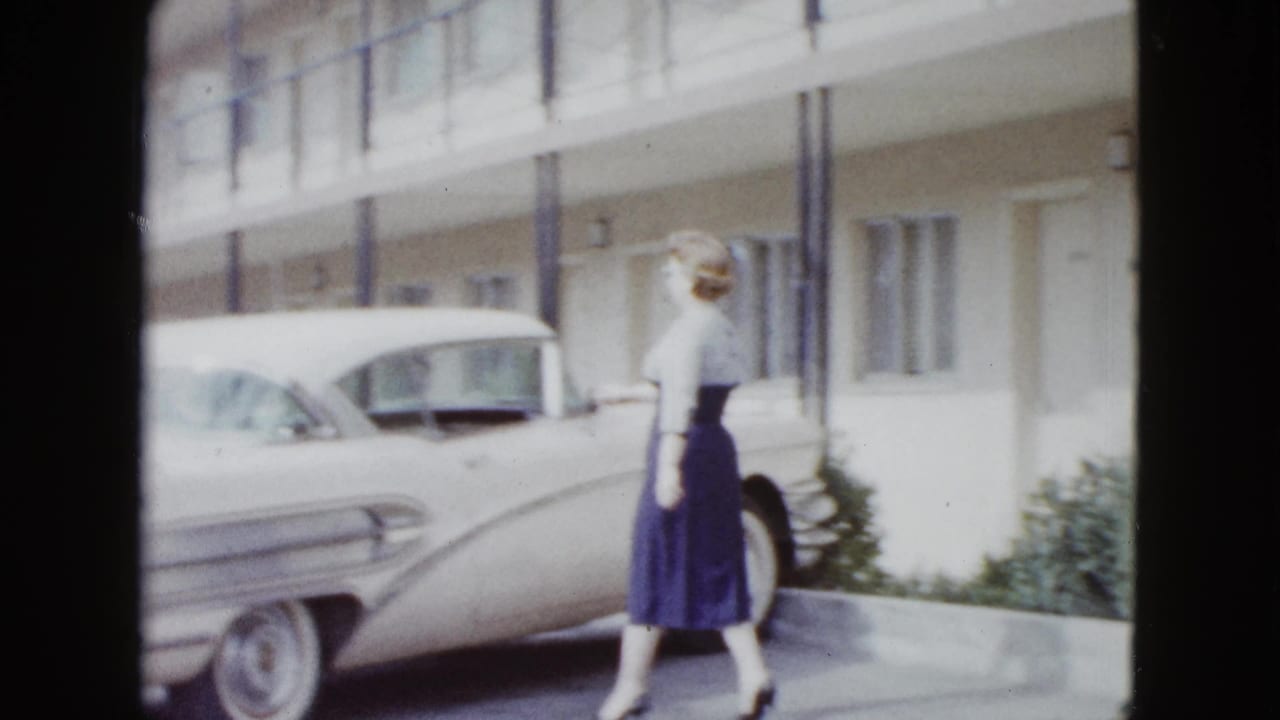 Woman in a blue dress walks beside a white classic car outside motel rooms.