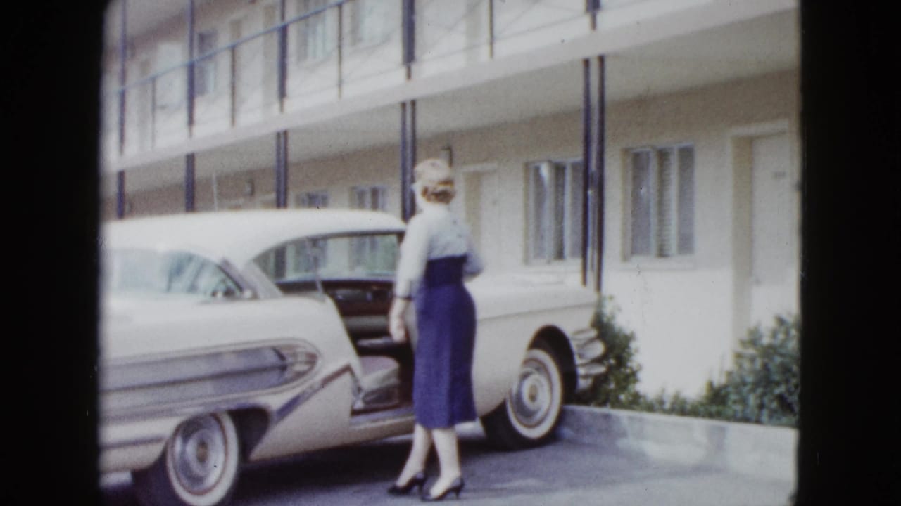 Woman stands near the open passenger door of a white car outside a motel.