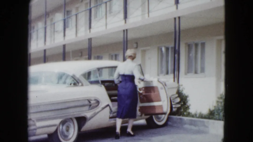 In supplied 1958 Las Vegas footage, the woman loads a brown suitcase through the open passenger door of the white classic car.