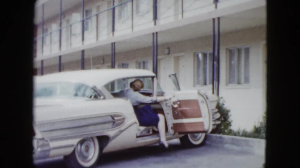 In supplied 1958 Las Vegas footage, the woman climbs into the passenger side of the white classic car outside motel-style lodging.