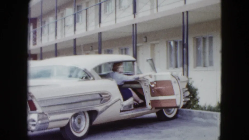 In 1958 Las Vegas footage, a woman sits by the open door of a white finned car parked outside a two-story motel.