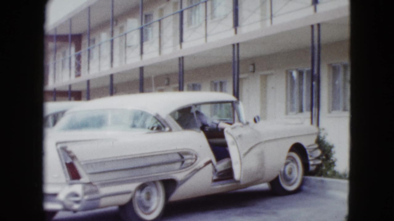 White late-1950s car with tail fins and an open door sits outside a Las Vegas motel corridor.