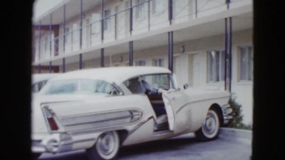 White late-1950s car with tail fins and an open door sits outside a Las Vegas motel corridor.