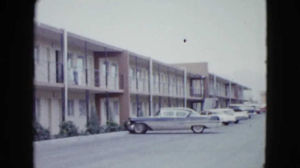 Motel exterior in Las Vegas with balcony railings, room doors, shrubs, and a parked 1950s car near the walkway.