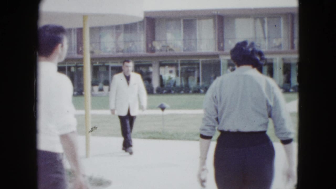 Man in a light jacket walks toward the camera as two adults move through a motel courtyard.